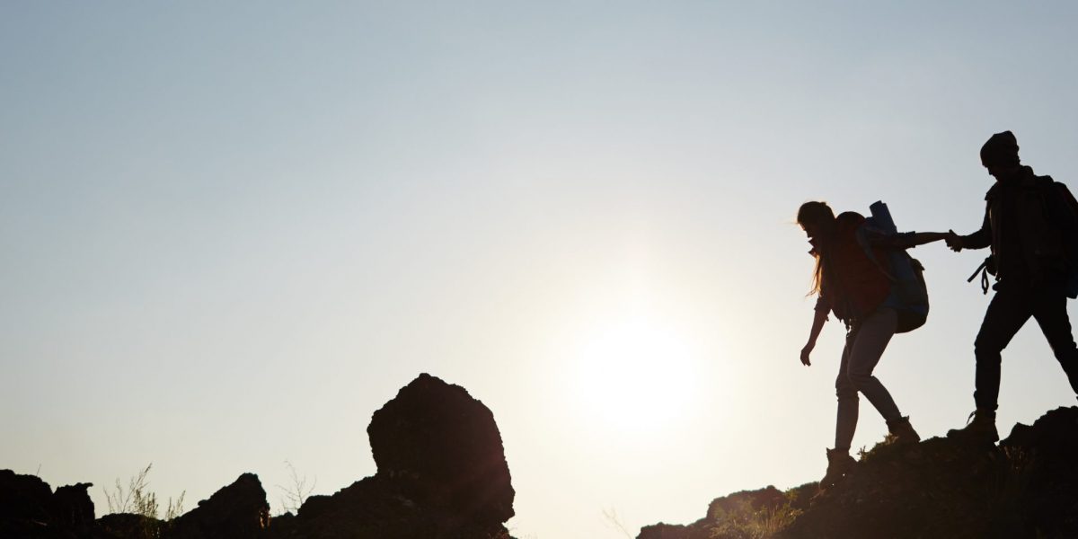Backlit dark silhouettes of couple hiking in mountains, young man following girlfriends holding hands, side view graphic image